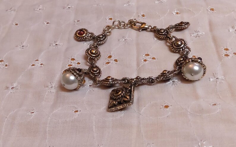 May include: Silver-toned bracelet with ornate detailing, featuring pearl and red gemstone accents. The bracelet has a decorative charm and is displayed on a white, patterned surface. The jewelry is a classic accessory.