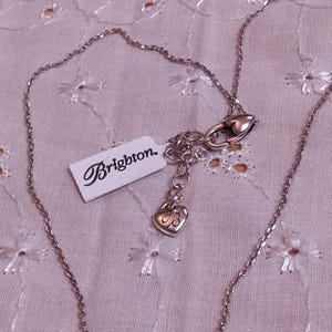 Op de afbeelding: Een zilverkleurige ketting met een fijne ketting en een hartvormige hanger. De ketting heeft een karabijnsluiting en een label met de tekst "Brighton". De sieraden worden gepresenteerd op een lichtroze, geborduurde stof.