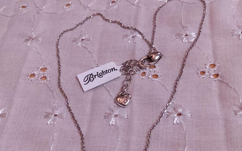 Op de afbeelding: Een zilverkleurige ketting met een fijne ketting en een hartvormige hanger. De ketting heeft een karabijnsluiting en een label met de tekst "Brighton". De sieraden worden gepresenteerd op een lichtroze, geborduurde stof.