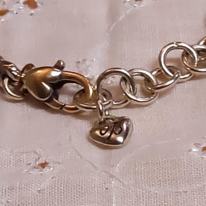 May include: Close-up of a silver-toned bracelet clasp with a gold-colored lobster claw clasp. A small heart-shaped charm with initials hangs from the clasp. The bracelet has a decorative, textured design.