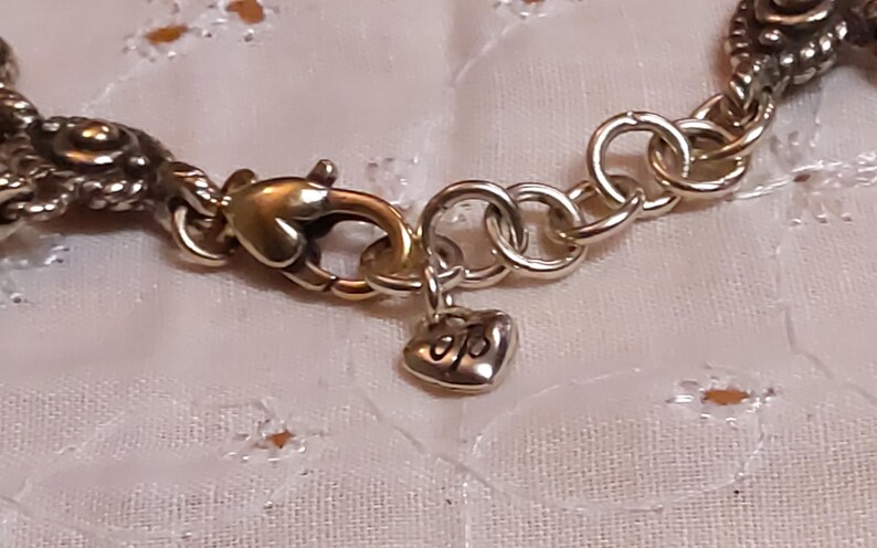 May include: Close-up of a silver-toned bracelet clasp with a gold-colored lobster claw clasp. A small heart-shaped charm with initials hangs from the clasp. The bracelet has a decorative, textured design.