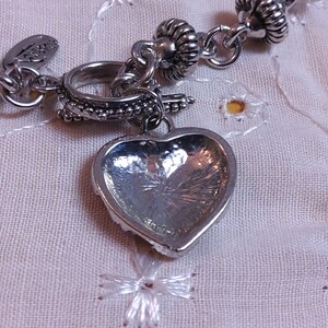 May include: A silver charm bracelet with a heart-shaped charm. The charm is detailed with a textured surface and a raised edge. The bracelet is made of a chain with small, round beads.