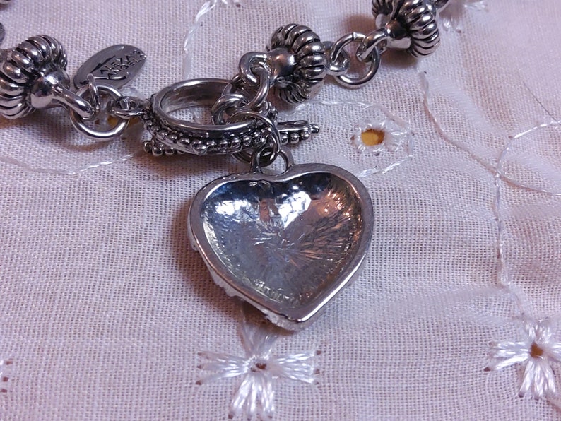 May include: A silver charm bracelet with a heart-shaped charm. The charm is detailed with a textured surface and a raised edge. The bracelet is made of a chain with small, round beads.