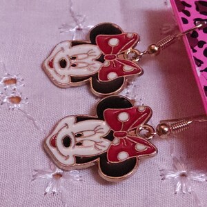 Pu&ograve; includere: Un paio di orecchini pendenti di Minnie Mouse. Ogni orecchino presenta un viso bianco, orecchie nere e un fiocco rosso a pois bianchi. Gli orecchini hanno ganci color oro e sono esposti su uno sfondo rosa.
