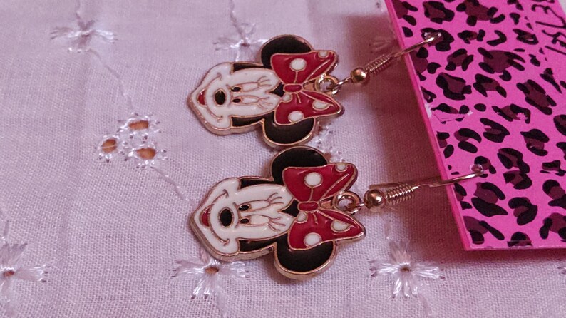 Pu&ograve; includere: Un paio di orecchini pendenti di Minnie Mouse. Ogni orecchino presenta un viso bianco, orecchie nere e un fiocco rosso a pois bianchi. Gli orecchini hanno ganci color oro e sono esposti su uno sfondo rosa.