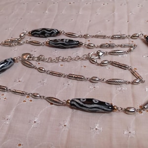Op de afbeelding: Een lange ketting met afwisselend zwart-wit patronen kralen en zilverkleurige metalen schakels. De ketting heeft ovale, cilindrische en bladvormige metalen elementen, met een karabijnsluiting. De ketting wordt gepresenteerd op een wit, getextureerd oppervlak.