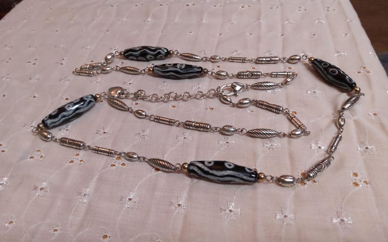 Op de afbeelding: Een lange ketting met afwisselend zwart-wit patronen kralen en zilverkleurige metalen schakels. De ketting heeft ovale, cilindrische en bladvormige metalen elementen, met een karabijnsluiting. De ketting wordt gepresenteerd op een wit, getextureerd oppervlak.