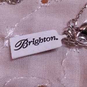 Op de afbeelding: Een zilverkleurige ketting met een decoratieve sluiting en een label met de tekst "Brighton". De ketting wordt gepresenteerd op een lichtroze, getextureerde stof.
