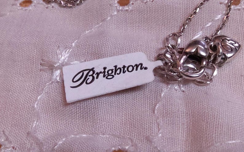 Op de afbeelding: Een zilverkleurige ketting met een decoratieve sluiting en een label met de tekst "Brighton". De ketting wordt gepresenteerd op een lichtroze, getextureerde stof.