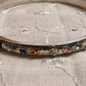 May include: Silver-toned bangle bracelet with a hammered texture. The bracelet is adorned with a variety of colorful gemstones, including purple, orange, pink, and blue. The gemstones are set in individual bezels around the bracelet.