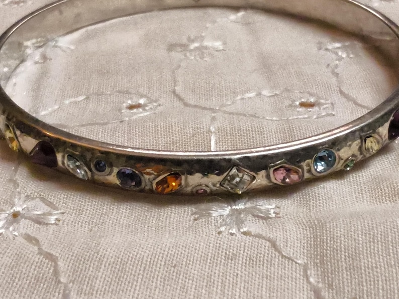May include: Silver-toned bangle bracelet with a hammered texture. The bracelet is adorned with a variety of colorful gemstones, including purple, orange, pink, and blue. The gemstones are set in individual bezels around the bracelet.