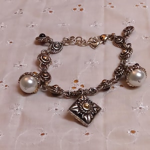 May include: Silver-toned bracelet with ornate detailing, featuring a square charm and pearl accents. The bracelet has a decorative design with gold-colored accents and a heart-shaped clasp. The bracelet is approximately 7 inches in length.