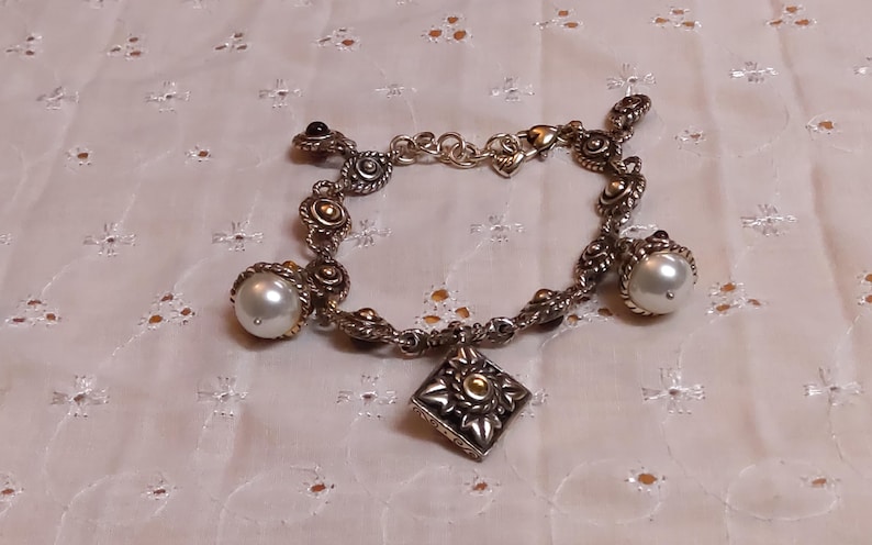 May include: Silver-toned bracelet with ornate detailing, featuring a square charm and pearl accents. The bracelet has a decorative design with gold-colored accents and a heart-shaped clasp. The bracelet is approximately 7 inches in length.