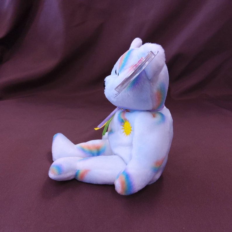 May include: A white plush rainbow bear with a yellow sun on its chest. The bear is sitting with its legs crossed and has a rainbow-colored tag attached to its ear.