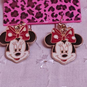 Pu&ograve; includere: Un paio di orecchini di Minnie Mouse dorati. Ogni orecchino presenta un viso bianco, orecchie nere e un fiocco rosso a pois bianchi. Gli orecchini sono su una carta rosa con stampa leopardata.