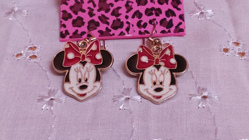 Pu&ograve; includere: Un paio di orecchini di Minnie Mouse dorati. Ogni orecchino presenta un viso bianco, orecchie nere e un fiocco rosso a pois bianchi. Gli orecchini sono su una carta rosa con stampa leopardata.