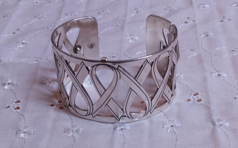 May include: Silver-toned cuff bracelet with an open design featuring interwoven ribbon-like shapes. The bracelet has a hinged opening for easy wear and is displayed on a white, patterned surface. The cuff is approximately 2 inches wide.