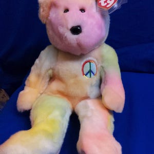 May include: A colorful plush teddy bear with a peace sign on its chest. The bear has a pink face and feet, with a rainbow-colored body. A heart-shaped tag is attached to the ear.