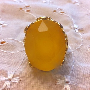 May include: A gold-toned ring featuring a large, oval-shaped, faceted yellow gemstone. The ring's setting has a decorative, scalloped edge. The ring is set against a white, embroidered fabric background.