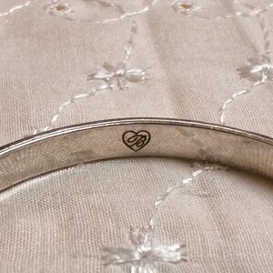 May include: A silver-toned bangle bracelet with a heart-shaped logo. The bracelet is smooth and curved, with a small heart and the letter "B" inside. The background is a light-colored, textured fabric.