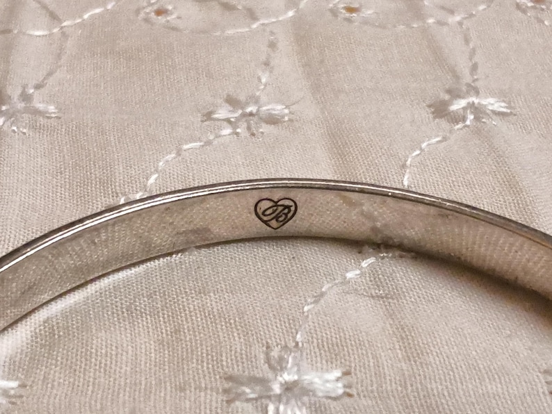 May include: A silver-toned bangle bracelet with a heart-shaped logo. The bracelet is smooth and curved, with a small heart and the letter "B" inside. The background is a light-colored, textured fabric.