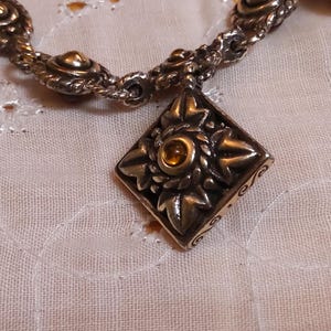 May include: Silver-toned bracelet with a decorative diamond-shaped pendant. The pendant features a central amber-colored stone surrounded by a floral design. The bracelet is on a white, textured surface.