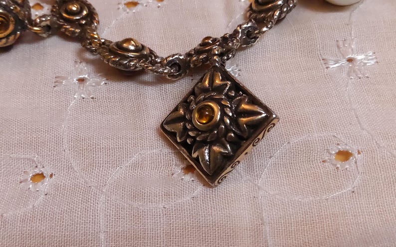 May include: Silver-toned bracelet with a decorative diamond-shaped pendant. The pendant features a central amber-colored stone surrounded by a floral design. The bracelet is on a white, textured surface.