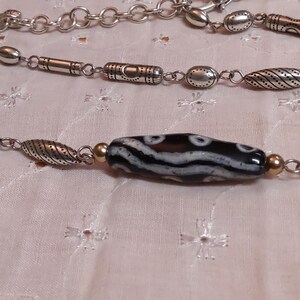 Op de afbeelding: Een zilverkleurige ketting met een centrale zwart-wit patroon kraal. De ketting heeft verschillende zilveren kralen en schakels. De achtergrond is een lichtroze stof met een bloemenpatroon.