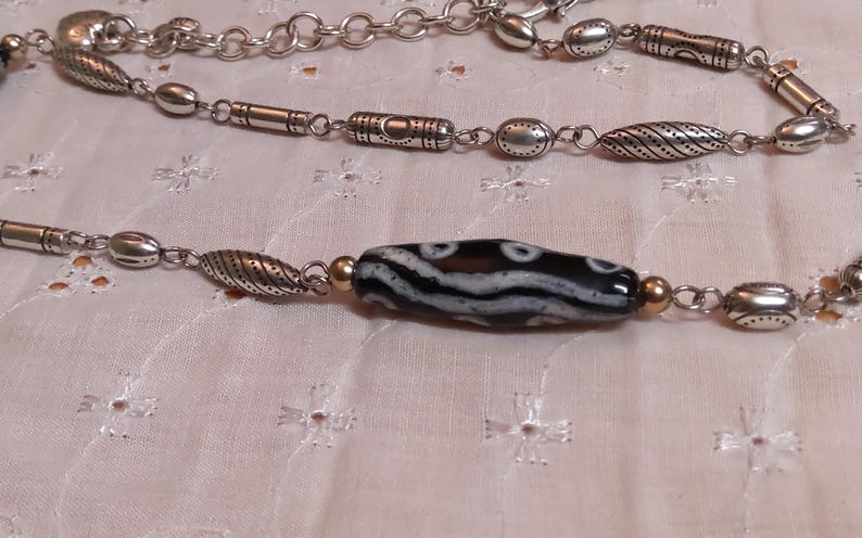 Op de afbeelding: Een zilverkleurige ketting met een centrale zwart-wit patroon kraal. De ketting heeft verschillende zilveren kralen en schakels. De achtergrond is een lichtroze stof met een bloemenpatroon.