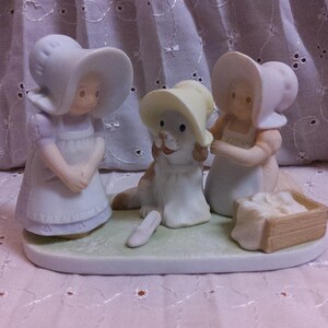 May include: A porcelain figurine featuring two girls in bonnets and aprons, flanking a dog wearing a bonnet. The dog is seated, and one girl holds a basket. The figurines are in shades of white, cream, and pale blue.