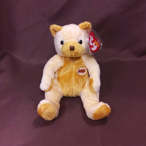 May include: A light yellow and brown plush teddy bear with a Ty Beanie Baby tag. The bear is sitting with its legs outstretched. The bear has a brown nose and black eyes.