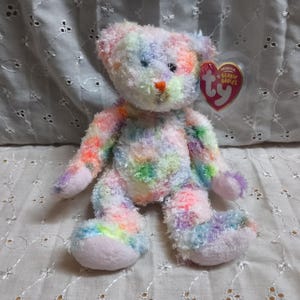 May include: A plush teddy bear featuring a multi-colored, fuzzy, tie-dye pattern in shades of pink, blue, yellow, and green. The bear has a small orange nose and a heart-shaped red tag attached to its ear with the text Ty Beanie Babies.