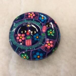 May include: A hand-painted, round decorative object with a dark blue base. The design features a central face with black eyes and a pink mouth, surrounded by pink, blue, and green floral patterns. The object is likely ceramic or pottery.