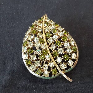 May include: A gold-tone leaf-shaped brooch with clear and green rhinestones.