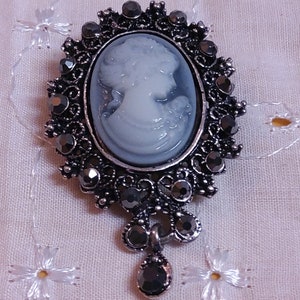May include: A silver cameo brooch with a white cameo of a woman's profile. The brooch is decorated with black rhinestones and has a dangling black rhinestone pendant.