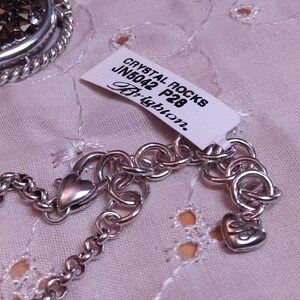 Może przedstawiać: Srebrny naszyjnik z łańcuszkiem i zawieszką w kształcie serca. Naszyjnik ma metkę z napisem "CRYSTAL ROCKS JN5042 P28 Brighton". Widoczny jest r&oacute;wnież okrągły wisiorek z ciemną, błyszczącą powierzchnią.