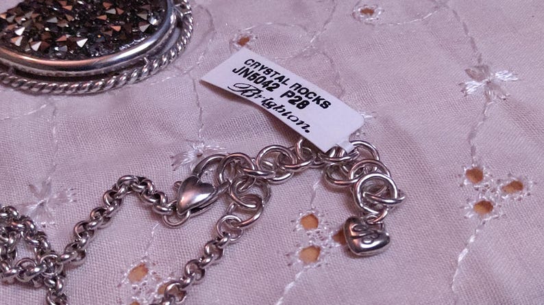 Może przedstawiać: Srebrny naszyjnik z łańcuszkiem i zawieszką w kształcie serca. Naszyjnik ma metkę z napisem "CRYSTAL ROCKS JN5042 P28 Brighton". Widoczny jest r&oacute;wnież okrągły wisiorek z ciemną, błyszczącą powierzchnią.