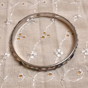 May include: A silver-toned bangle bracelet with a decorative band featuring small, colorful stones. The bracelet is circular and rests on a cream-colored fabric with a floral pattern. The bangle is a piece of jewelry.