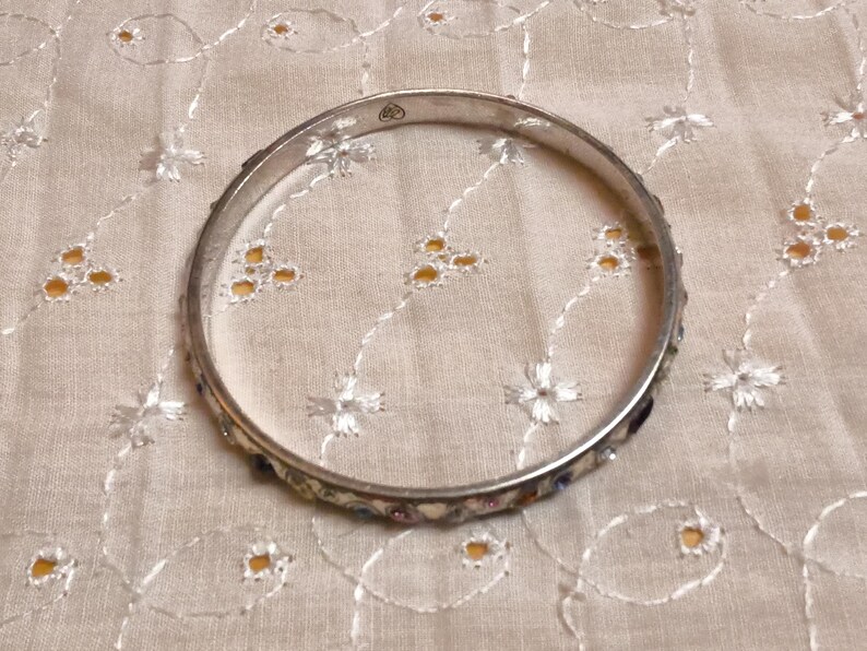 May include: A silver-toned bangle bracelet with a decorative band featuring small, colorful stones. The bracelet is circular and rests on a cream-colored fabric with a floral pattern. The bangle is a piece of jewelry.