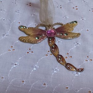 May include: A decorative dragonfly ornament with gold-toned wings and a pink center stone. The wings are accented with green and clear crystals. A sheer ribbon is attached for hanging.