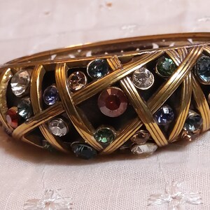 May include: A vintage gold-toned bangle bracelet with a woven design. The bracelet is adorned with a variety of colorful gemstones, including red, blue, green, and clear crystals. The bracelet is approximately 2 inches wide.
