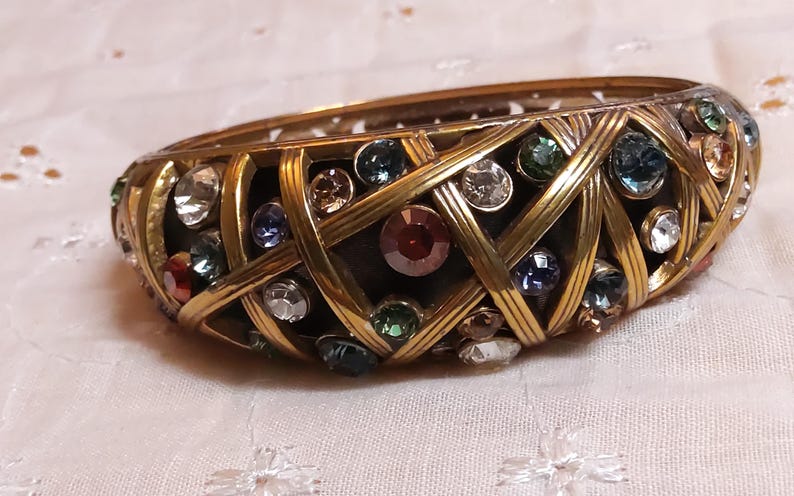 May include: A vintage gold-toned bangle bracelet with a woven design. The bracelet is adorned with a variety of colorful gemstones, including red, blue, green, and clear crystals. The bracelet is approximately 2 inches wide.