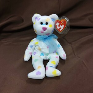 May include: White plush teddy bear with a blue ribbon around its neck. The bear is covered in colorful heart-shaped spots. The bear has a Ty Beanie Babies tag attached to its ear.