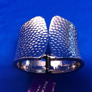 May include: Silver-toned cuff bracelet with a textured, hammered metal design. The bracelet has a hinged opening and a clasp closure. The surface is covered in small, evenly spaced dimples. The bracelet is displayed on a blue surface.