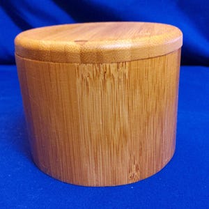 May include: A cylindrical wooden container with a lid. The container and lid are made of light brown bamboo. The container is sitting on a blue surface.