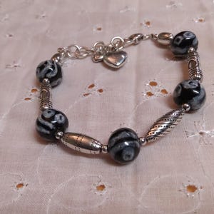 May include: A beaded bracelet featuring black and white patterned beads interspersed with silver-toned metal beads and accents. The bracelet includes a small heart charm and is displayed on a white, textured surface.