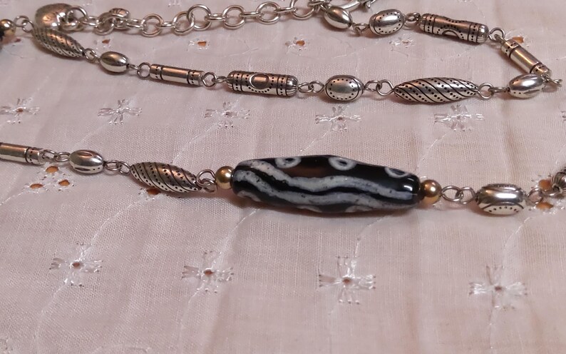 Op de afbeelding: Een zilveren ketting met verschillende kraalvormen en een centrale zwart-wit patroon kraal. De ketting heeft ovale, cilindrische en getextureerde kralen, verbonden door kleine schakels. De achtergrond is een lichtgekleurde stof.