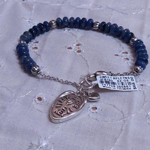 Puede incluir: Una pulsera de cuentas azules con un dije plateado. La pulsera presenta peque&ntilde;as cuentas azul oscuro y detalles plateados. Un dije plateado con un dise&ntilde;o grabado cuelga de la pulsera. Una etiqueta con texto est&aacute; adjunta.