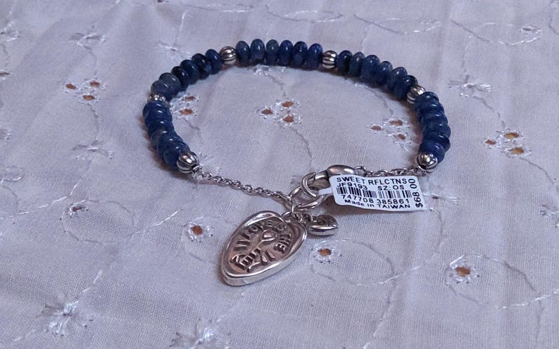 Puede incluir: Una pulsera de cuentas azules con un dije plateado. La pulsera presenta peque&ntilde;as cuentas azul oscuro y detalles plateados. Un dije plateado con un dise&ntilde;o grabado cuelga de la pulsera. Una etiqueta con texto est&aacute; adjunta.