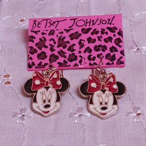 Pu&ograve; includere: Orecchini di Minnie Mouse con fiocco rosso e viso bianco, appesi a un cartoncino rosa con "Betsey Johnson" e stampa leopardata. Un accessorio divertente.